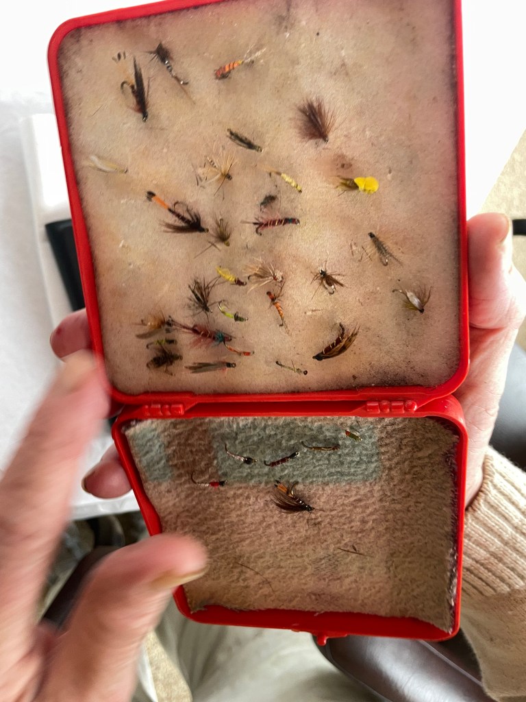 an elederly man's  hand holding a box of hand tied fishing flies